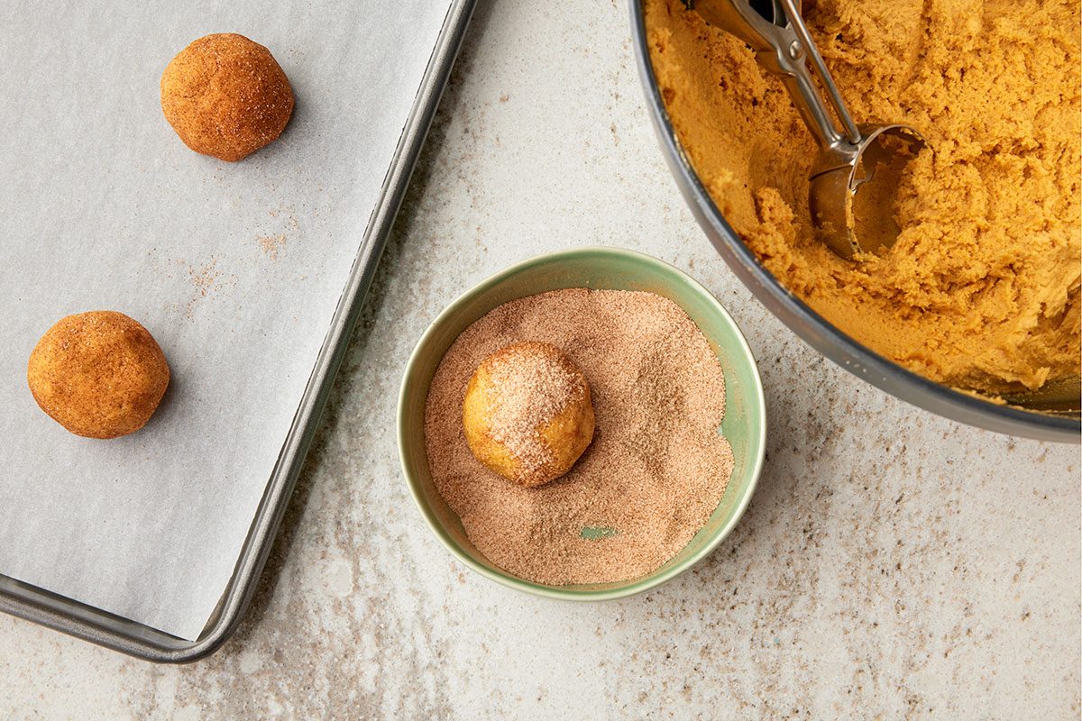 Overhead shot of cookie dough rolled in cinnamon sugar; with a tray of dough balls and a mixing bowl with more dough nearby; all set on a light countertop;