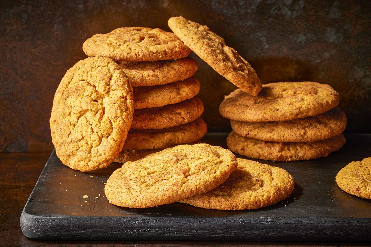 Close shot of pumpkin snickerdoodles, stacked and scattered on a dark slate surface; with golden-brown tops coated in cinnamon sugar;
