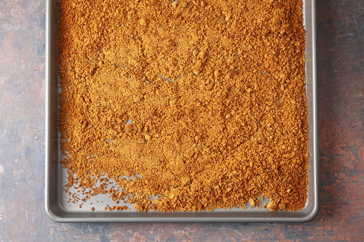 A metal baking sheet holds an even layer of crumbled, golden-brown toasted mixture, spread out thinly and covering most of the surface.