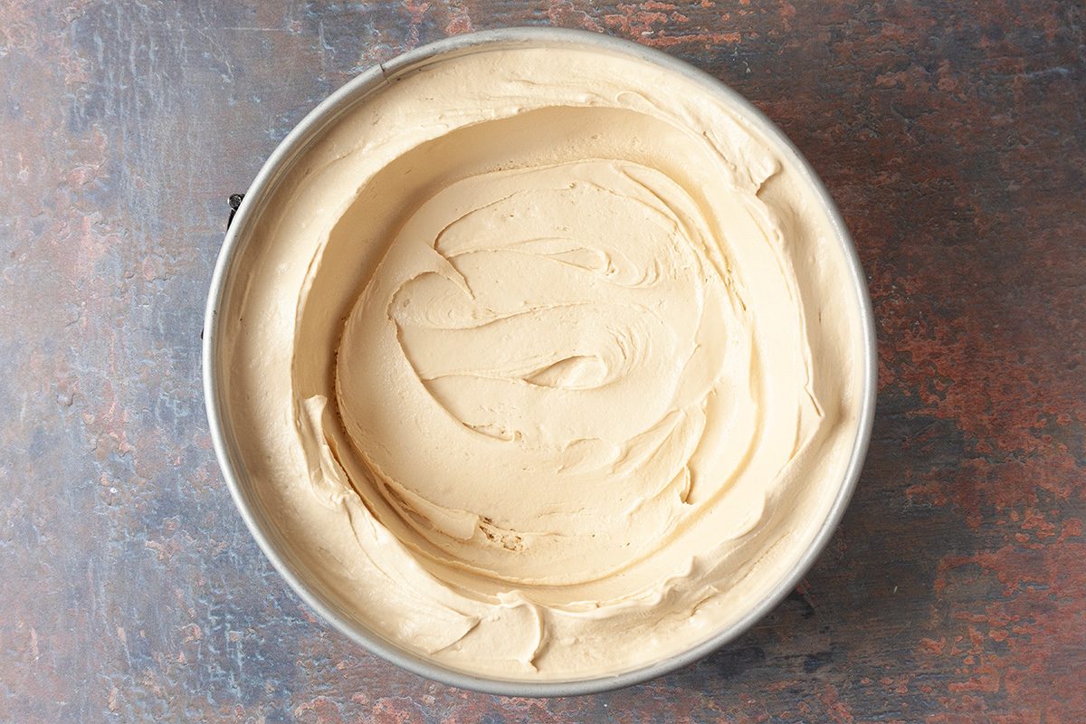 A round metal cake pan filled with smooth, light brown batter is placed on a rustic brown surface. The batter is evenly spread with visible swirls on the top.