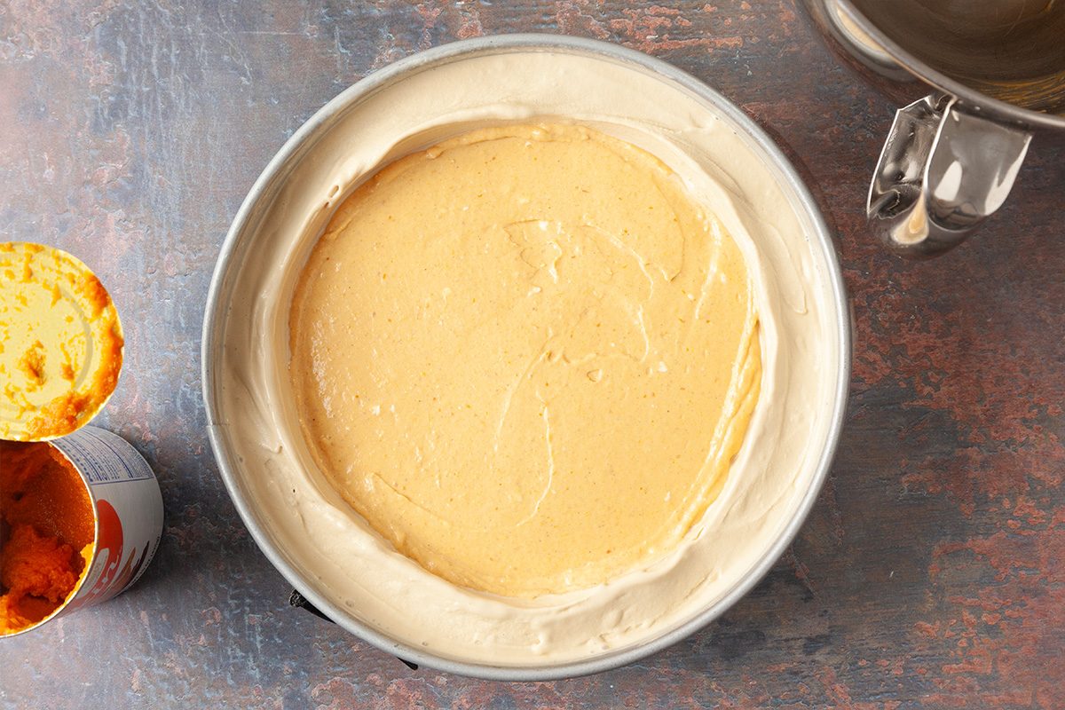 A round cake pan filled with a creamy, light brown batter sits on a textured surface. An empty mixing bowl and an opened can with orange residue are beside it.
