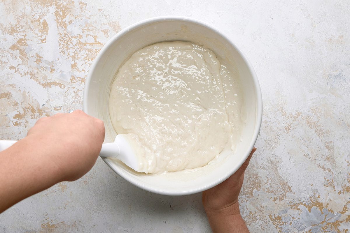 A person uses a white spatula to mix a thick, pale batter in a large white bowl on a lightly textured surface.