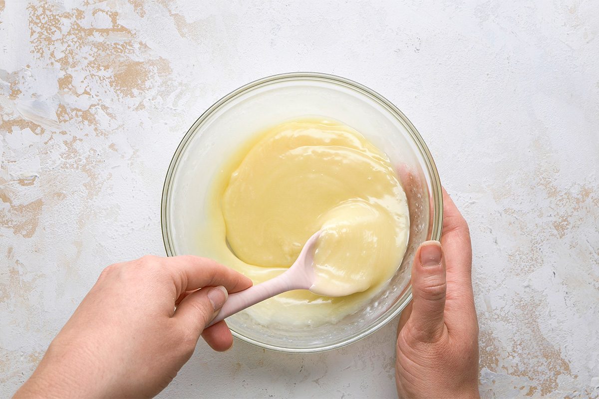 A person stirs a creamy, pale yellow mixture in a glass bowl with a light pink spatula on a light textured surface.