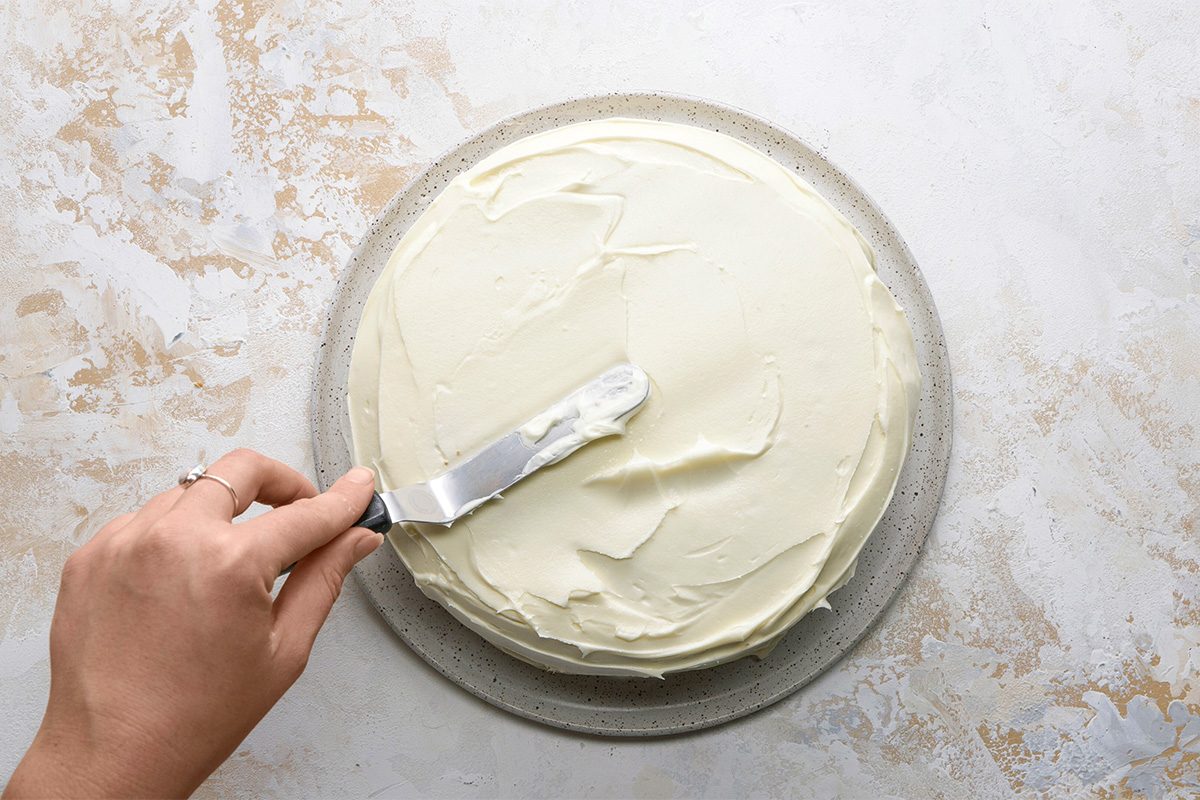 A hand using an offset spatula to spread smooth white frosting over a round cake on a speckled plate, set against a light textured background.