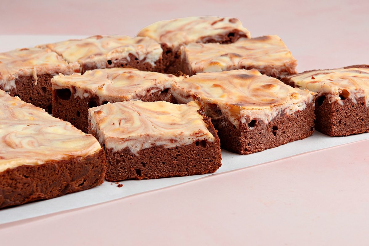 Squares of chocolate brownies with a creamy, swirled topping are arranged on a white surface against a light pink background.