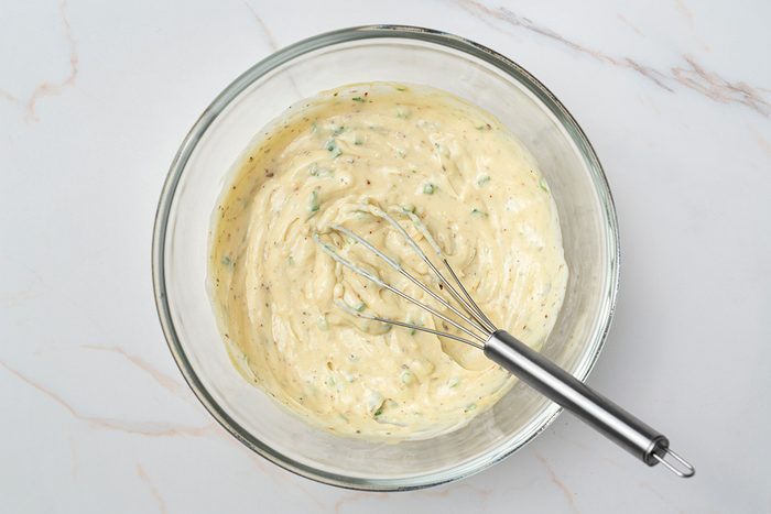 A glass bowl filled with a creamy, herb-speckled sauce sits on a white marble surface, with a metal whisk resting in the bowl.