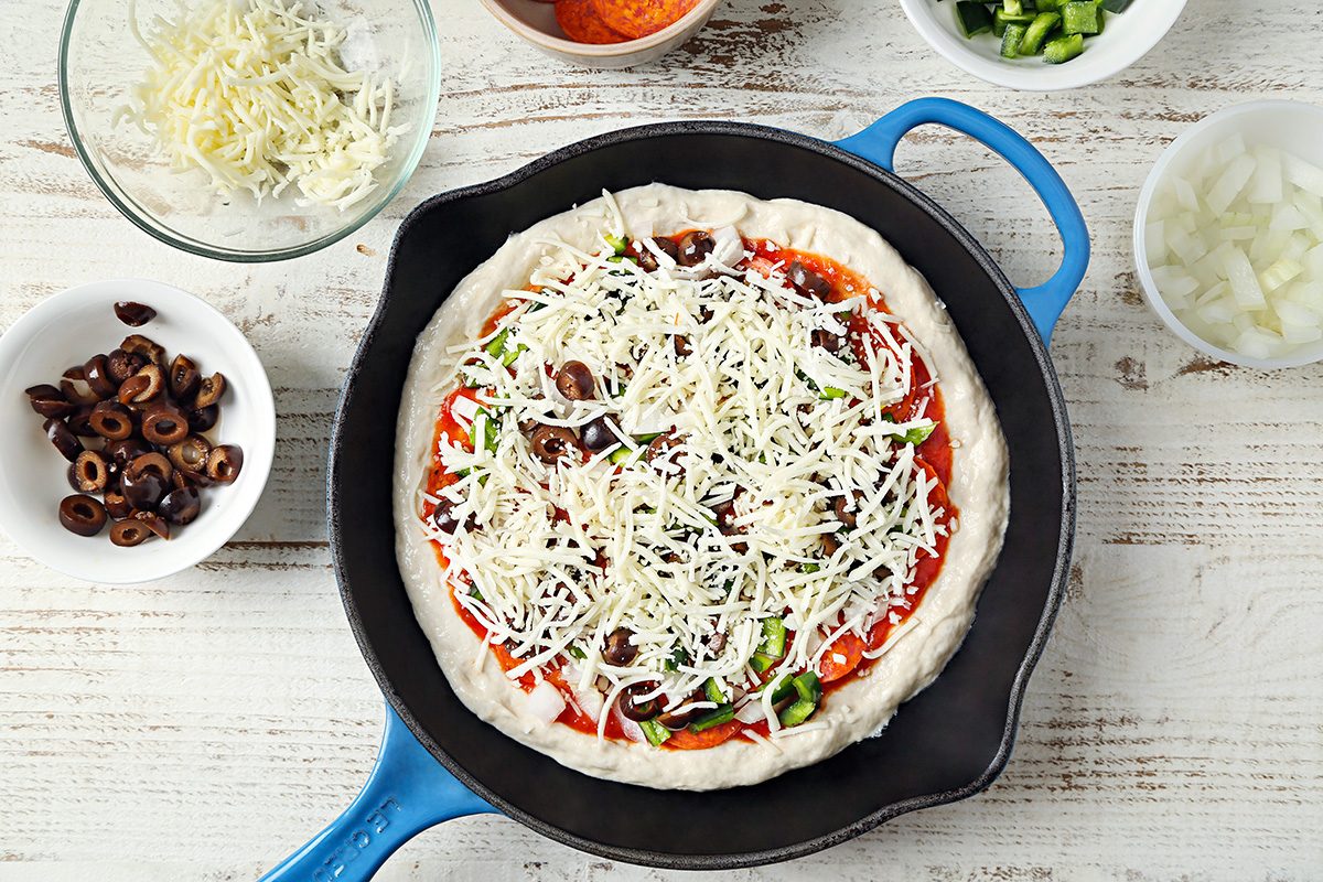 A cast iron skillet holds an uncooked pizza topped with cheese, olives, and vegetables. Bowls of shredded cheese, sliced olives, chopped onions, and green peppers surround it on a white wooden surface.