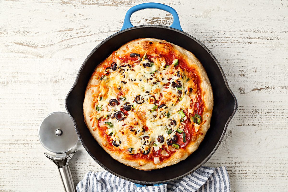 A homemade pizza with melted cheese, olives, and vegetables in a black skillet, next to a pizza cutter and a striped kitchen towel on a white wooden surface.