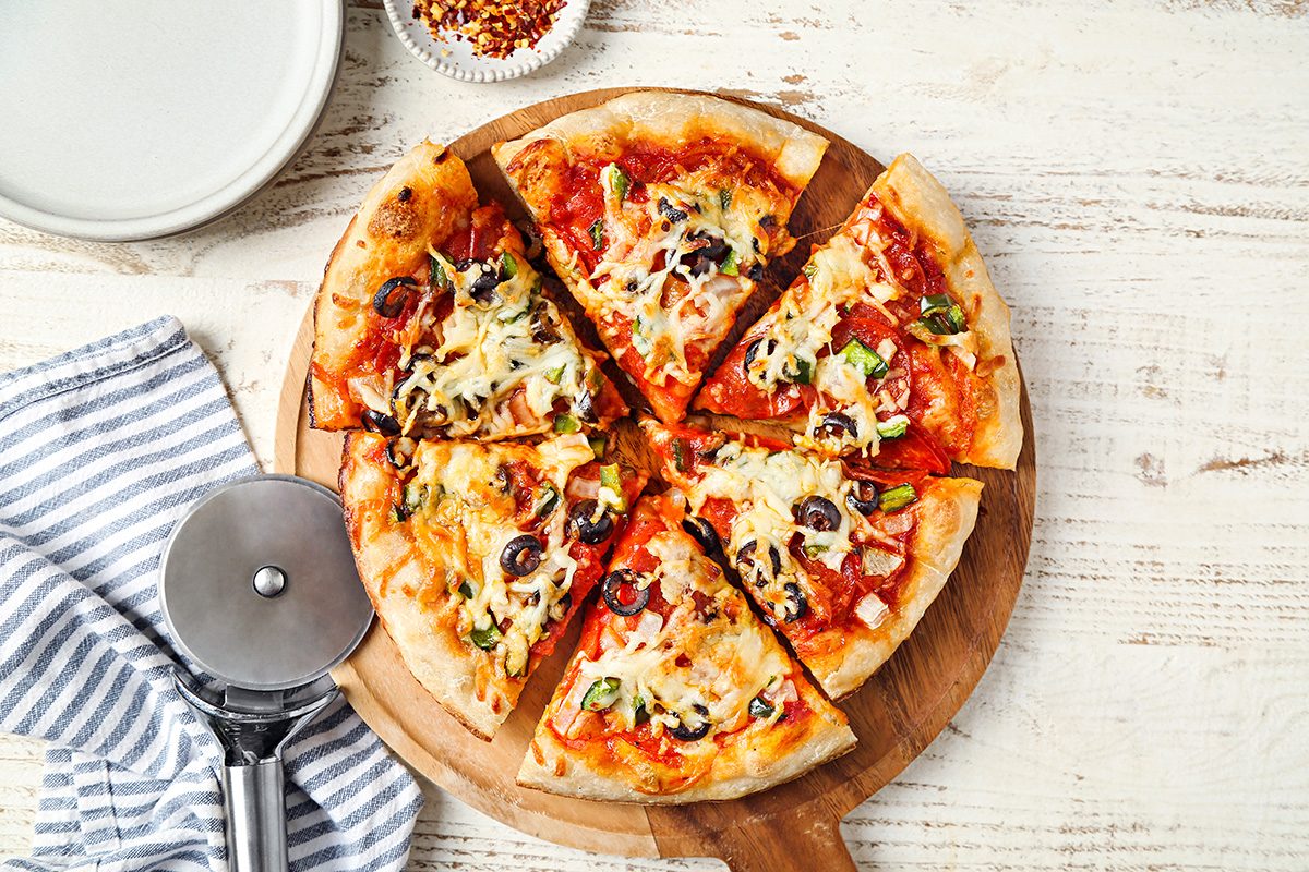 A sliced pizza with cheese, olives, and vegetables sits on a wooden board next to a pizza cutter, striped napkin, and small bowl of red pepper flakes on a light wooden table.