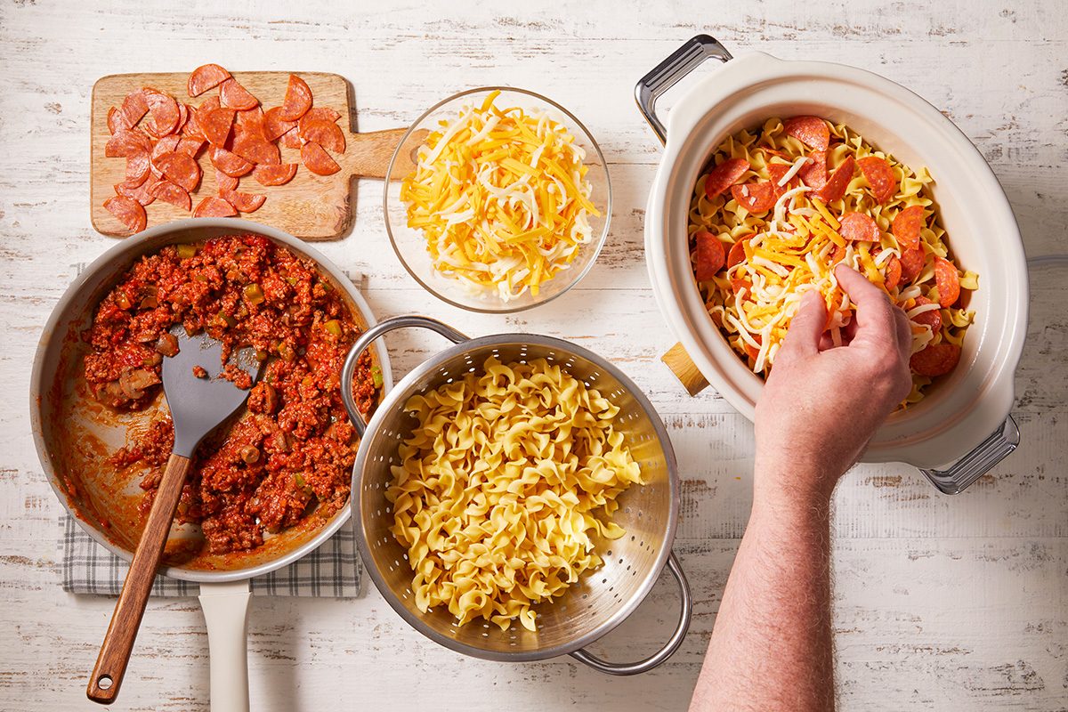 A person assembles a pasta bake, layering cooked pasta, ground meat sauce, shredded cheese, and sliced pepperoni in a baking dish, with ingredients and utensils arranged on a white wooden surface.