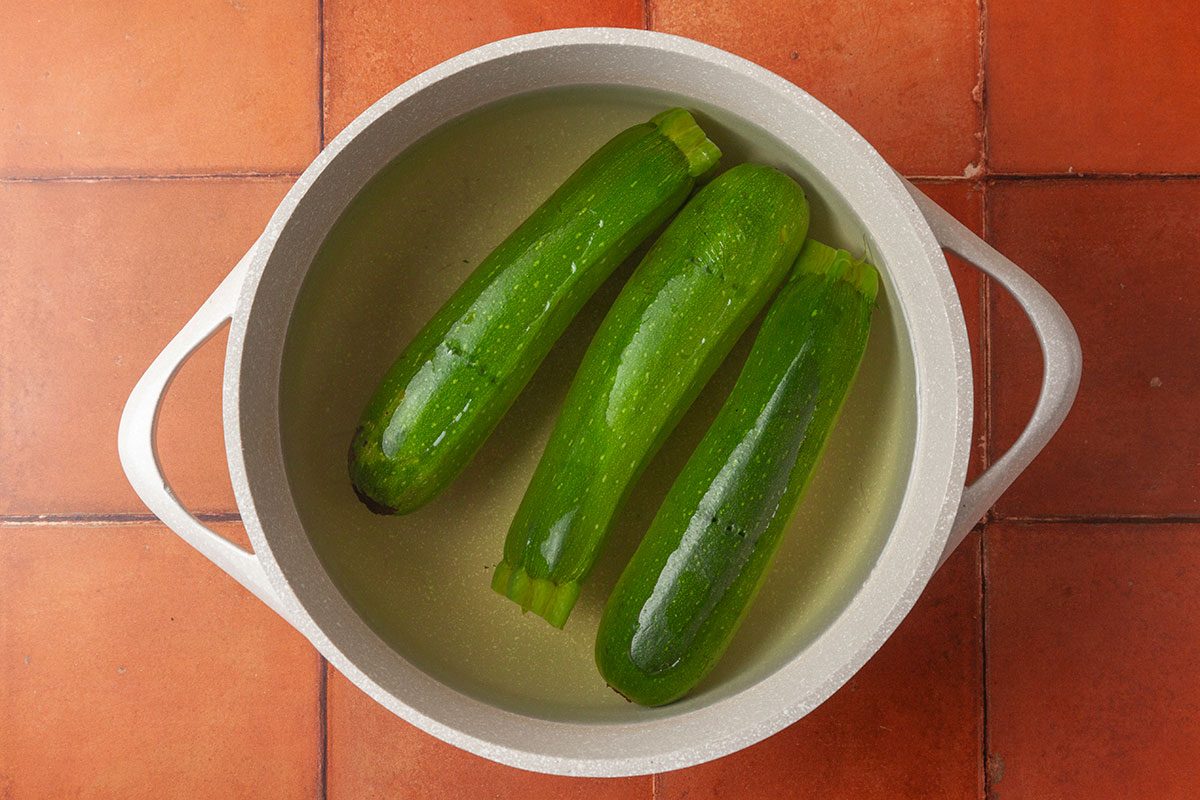 Overhead shot of three whole zucchinis soaking in water inside a light-colored pot; set on a terracotta-tiled surface