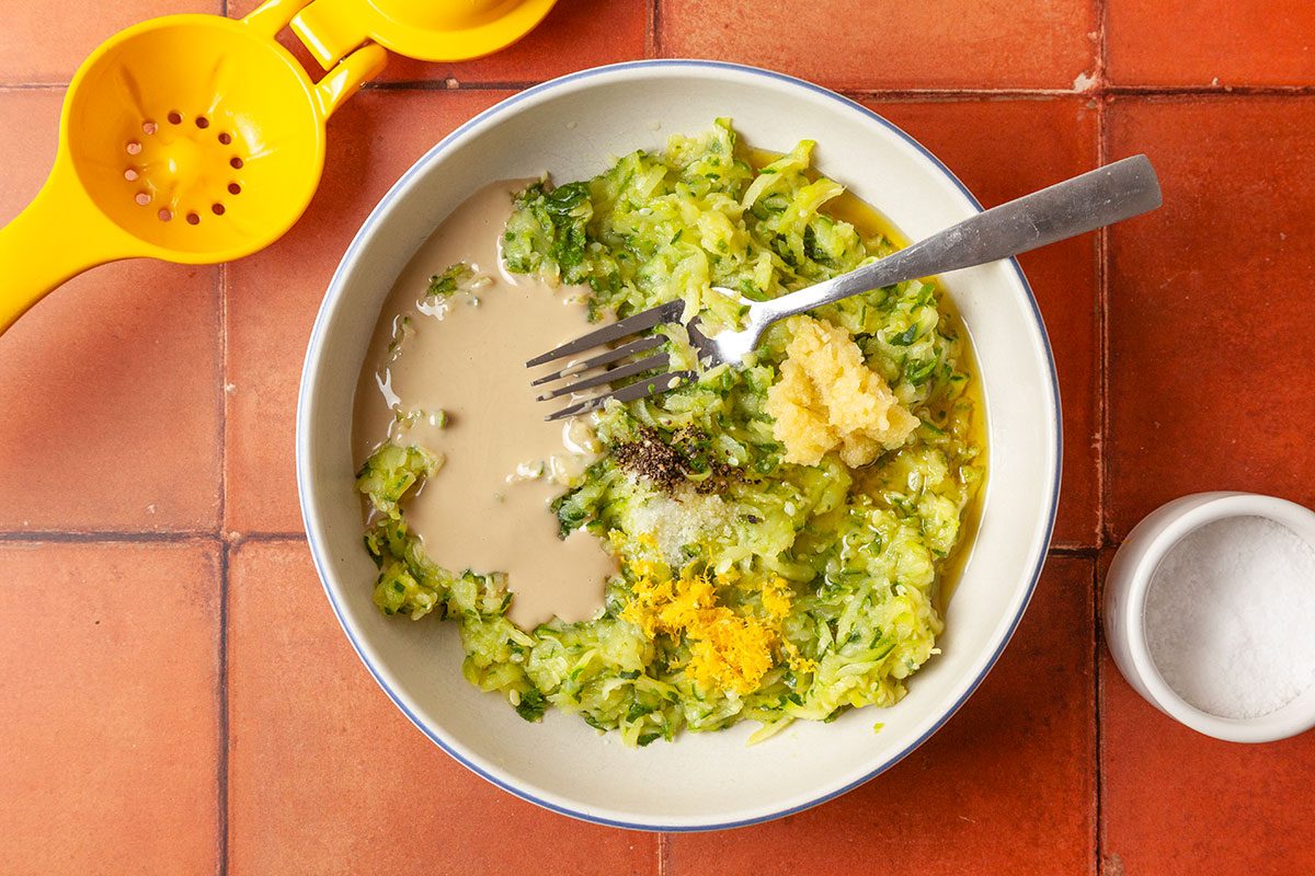 Overhead shot of a bowl with shredded zucchini, tahini, minced garlic, lemon zest; and black pepper with a fork; alongside a yellow citrus squeezer and a small bowl of salt; set on a terracotta-tiled surface