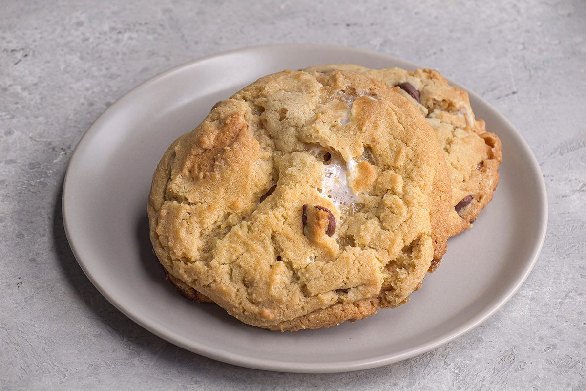 Two large chocolate chip cookies are stacked on a light gray plate, placed on a textured gray surface. The cookies look soft and slightly golden brown.