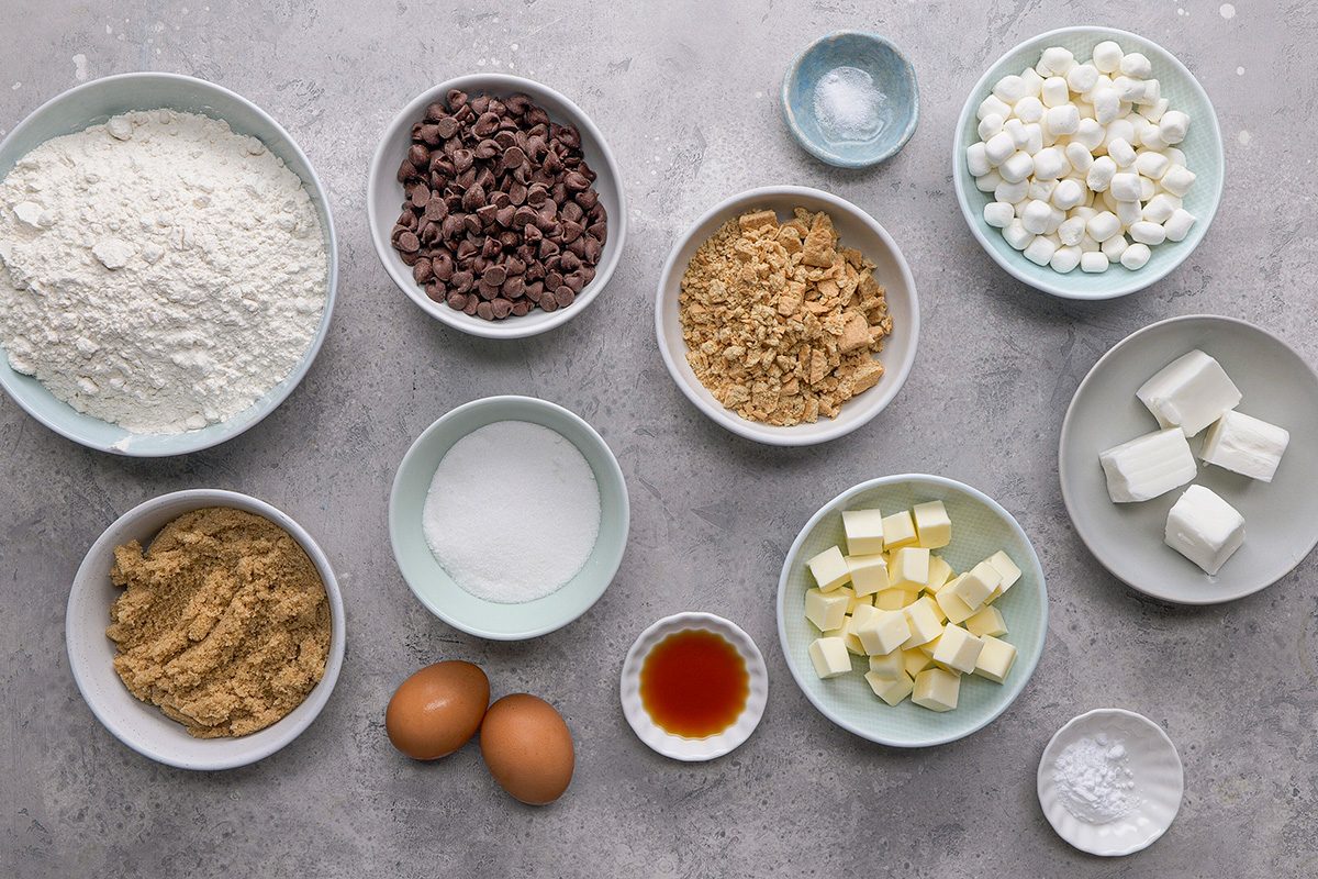 Various baking ingredients are arranged in bowls on a gray surface, including flour, chocolate chips, brown sugar, white sugar, mini marshmallows, eggs, butter cubes, vanilla extract, cream cheese, and baking powder.