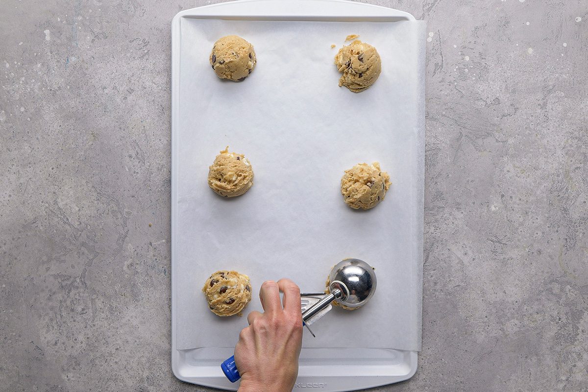 A hand uses a cookie scoop to place cookie dough balls on a baking sheet lined with parchment paper; five dough balls are already spaced on the sheet.