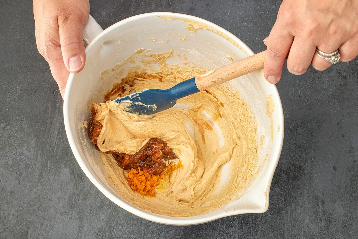 Overhead shot of hands holding a white mixing bowl filled with light brown batter; as a blue spatula stirs in dark brown and orange ingredients, all set on a black surface