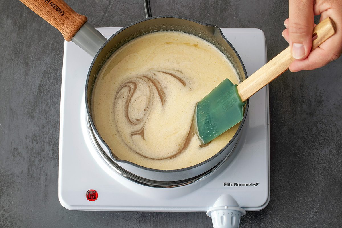 Overhead shot of a hand stirring a creamy mixture with a green spatula in a saucepan on a white electric stove; with visible swirls in the liquid