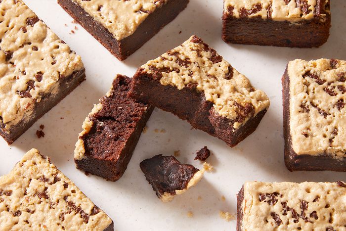 Squares of chocolate brownies topped with a layer of cookie dough are arranged on a light surface; one brownie in the center has a bite taken out, showing its fudgy texture.