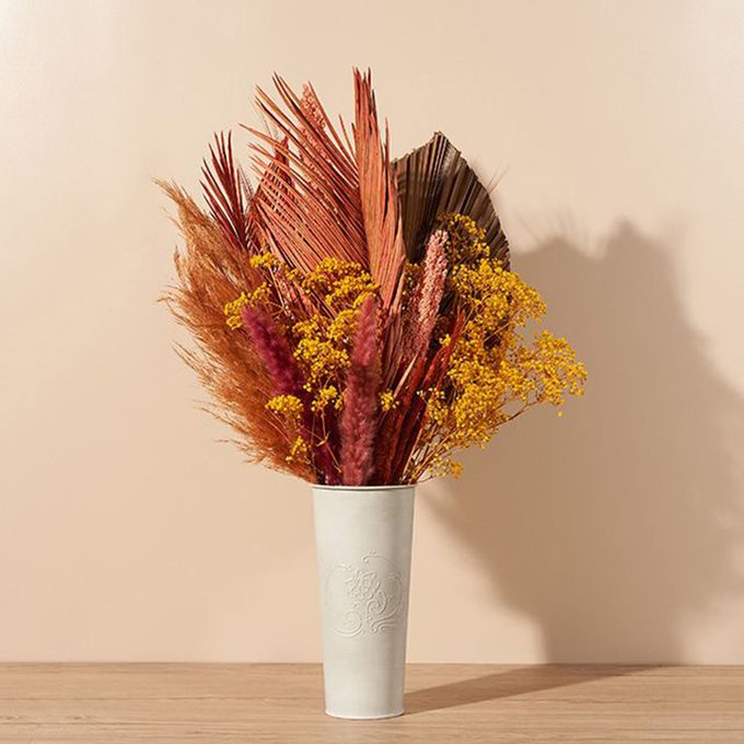 A white textured vase holds an arrangement of dried orange, brown, and yellow flowers and foliage, set against a beige background on a wooden surface.