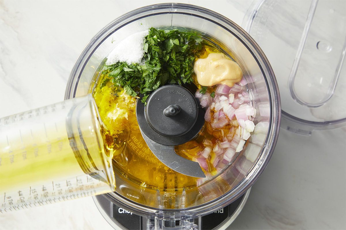 A food processor bowl containing chopped herbs, diced red onions, a dollop of mayonnaise, and olive oil, ready to be blended.