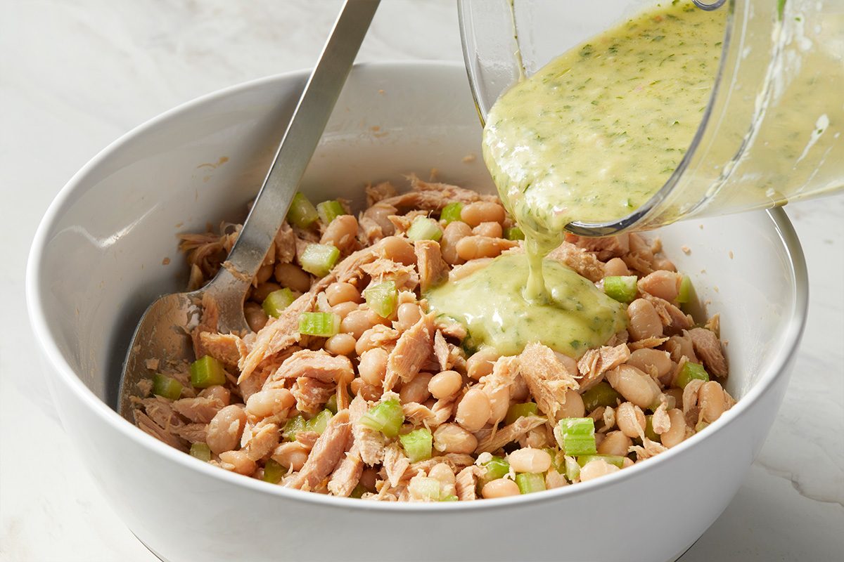 A bowl filled with flaked tuna, white beans, and chopped green onions, with a creamy green dressing being poured in from a blender. A metal spoon rests in the bowl.