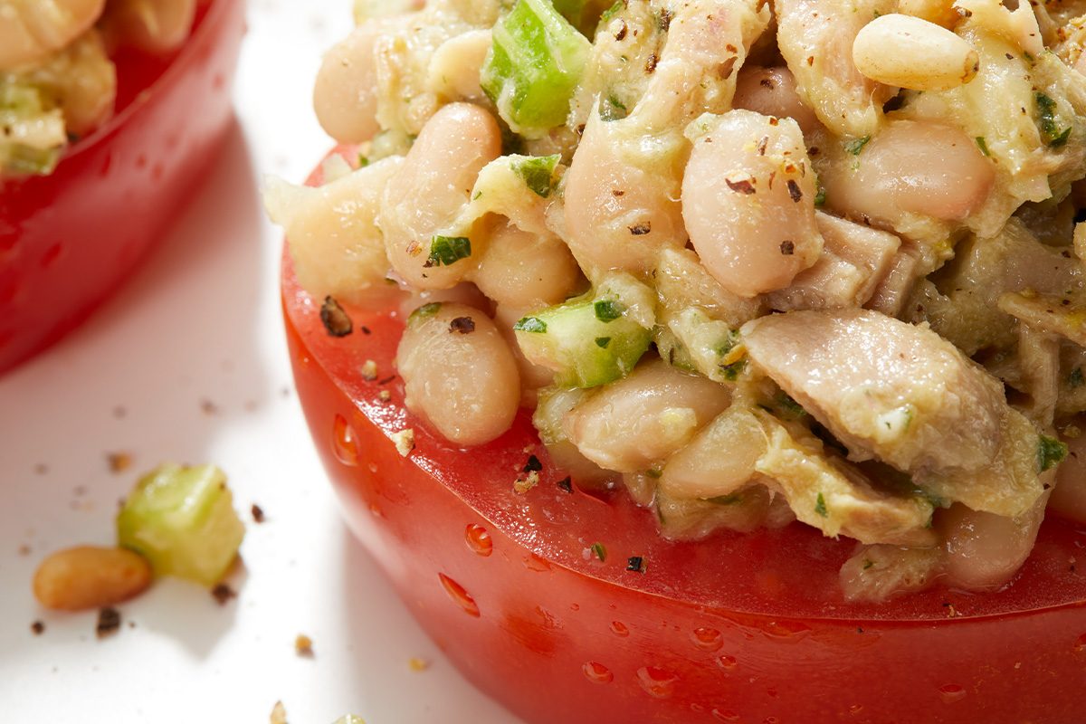 Close-up of a halved tomato topped with a mixture of white beans, diced celery, herbs, and black pepper. The fresh ingredients are clearly visible and the dish appears vibrant and healthy.