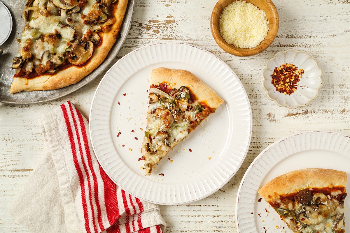 Overhead view of two slices of mushroom pizza on white plates, with a larger partial pizza, a bowl of grated cheese, a dish of red pepper flakes, and a red-striped napkin on a white wooden table.