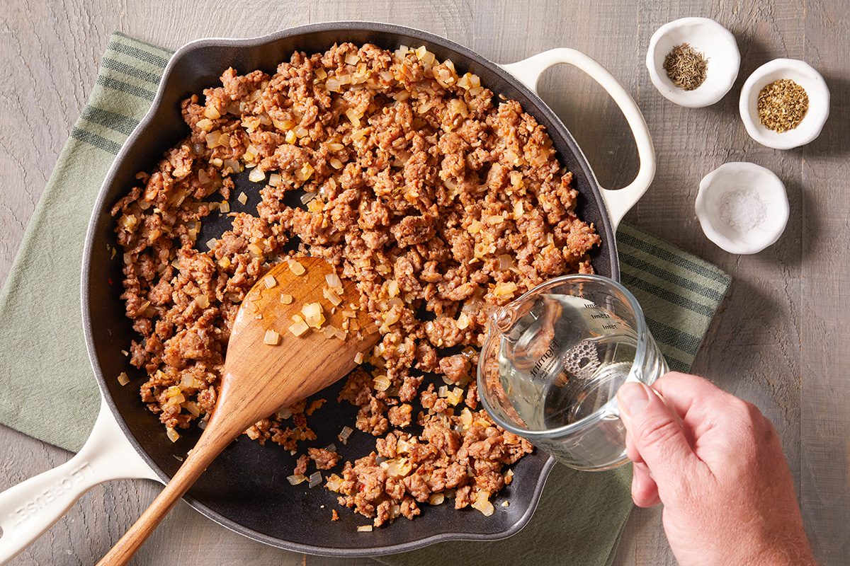 A hand pours water into a skillet with browned ground meat and onions, stirred with a wooden spoon. Two small bowls with spices are on the table beside a green-striped cloth.