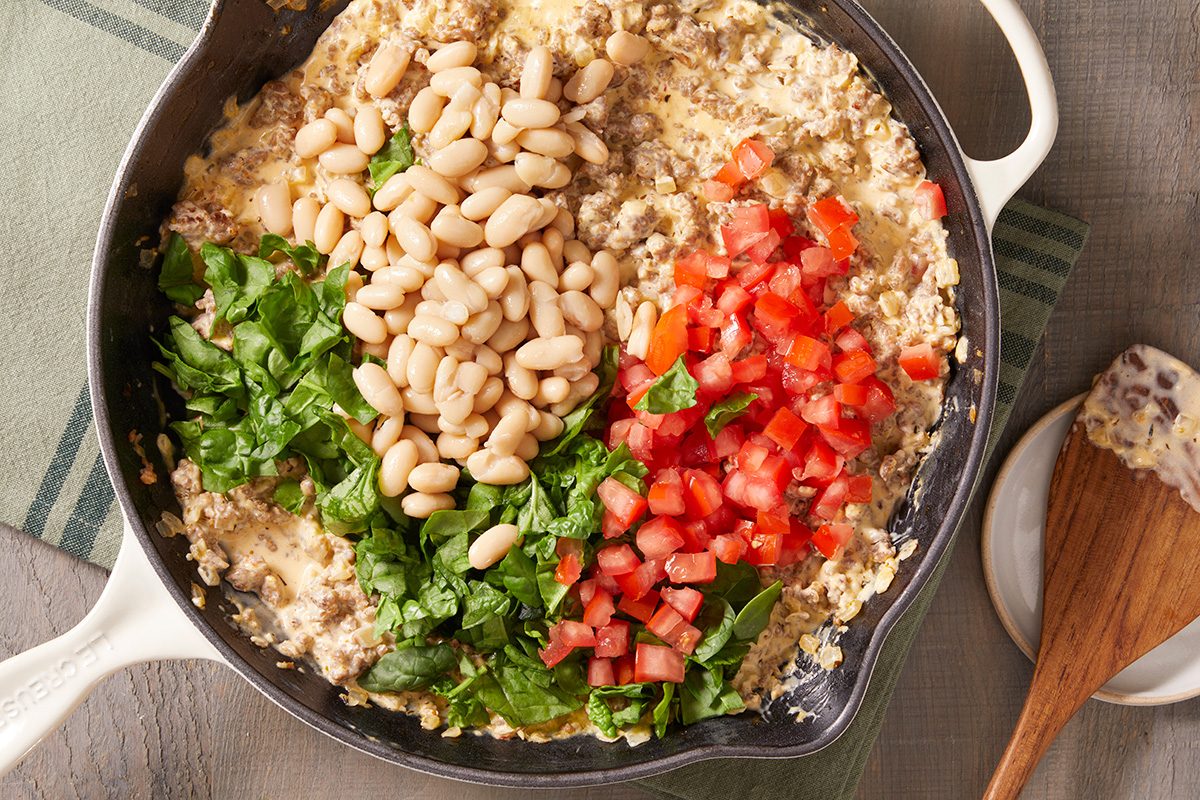 A skillet filled with a mixture of oatmeal, white beans, chopped spinach, and diced tomatoes, seen from above. A wooden spatula rests on a nearby plate.
