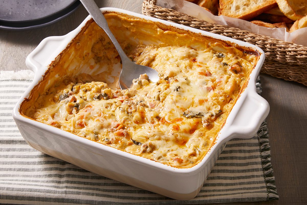 A creamy baked casserole in a white square dish with a serving spoon, showing a portion already scooped out. The dish rests on a striped cloth next to a basket of sliced bread.