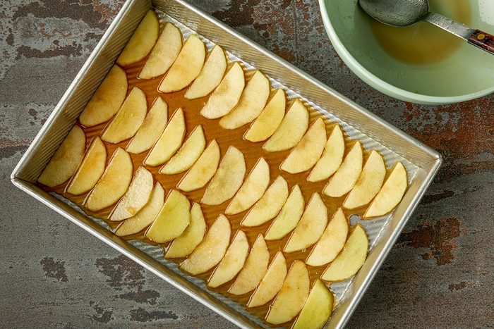 Overhead shot of a rectangular baking pan filled with neatly arranged apple slices on a layer of syrup; placed beside a green bowl containing liquid and a spoon