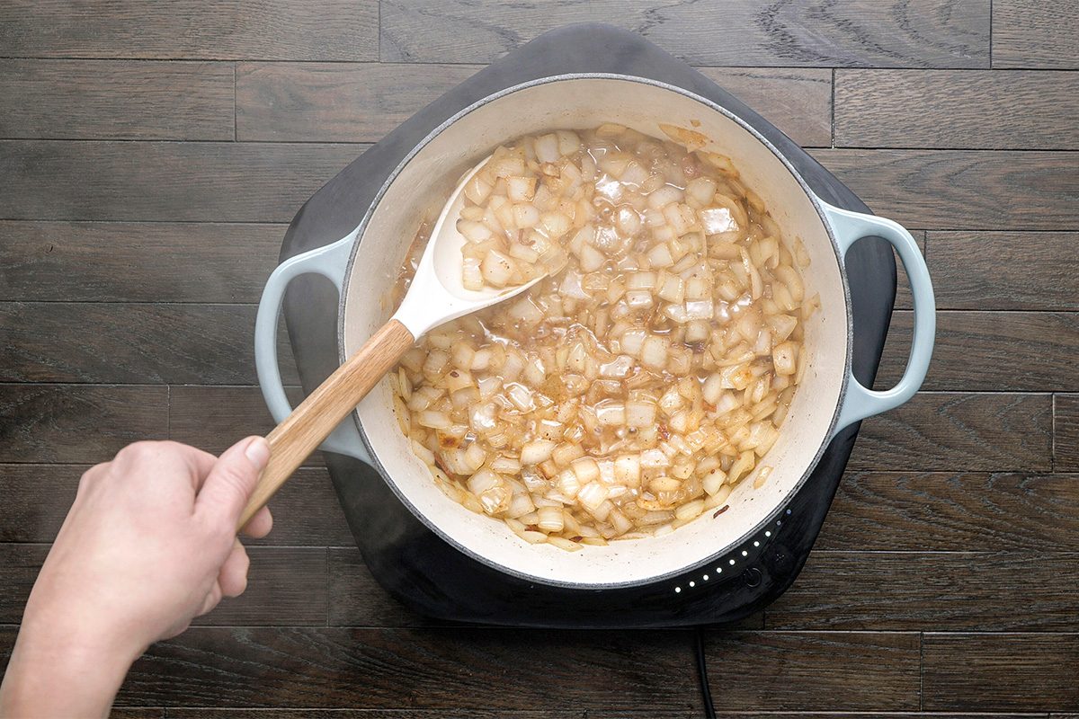 A hand stirs chopped onions cooking in a white pot on a stovetop, placed on a wooden surface. The onions appear to be sautéing and turning translucent.