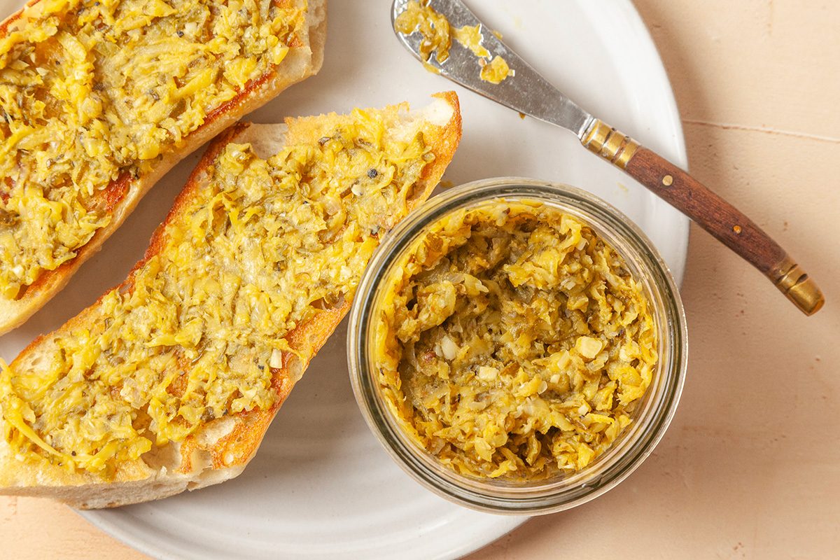 This is a close-up shot; a jar of artichoke spread and toasted bread topped with chunky yellow-green spread sit on a white plate; a wooden-handled knife lies beside them; the background is minimal and clean