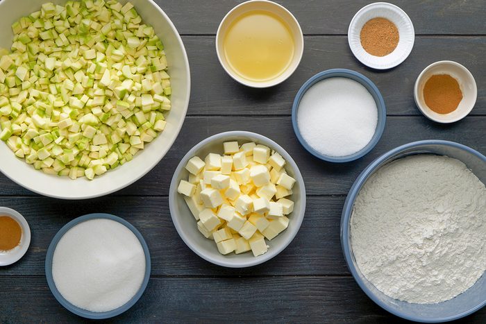 Several bowls containing recipe ingredients sit on a dark wooden surface, including chopped green apples, cubed butter, flour, sugar, liquid, and various spices measured out in small dishes.