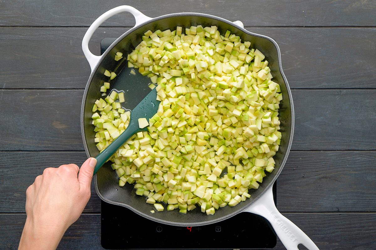 A hand stirs chopped celery and onions in a large white skillet with a green spatula on a stovetop.