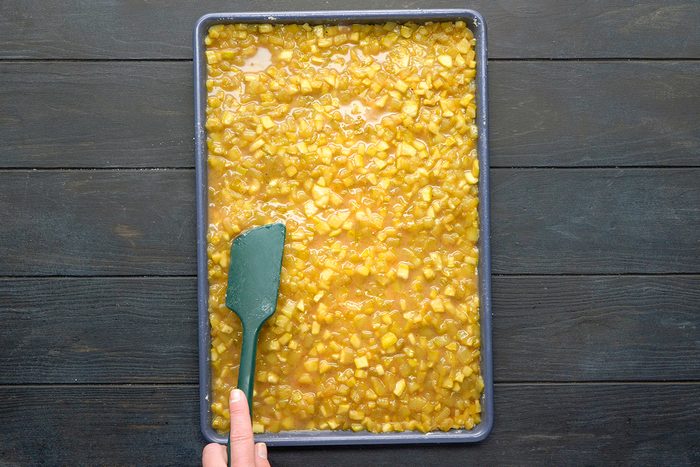 A hand uses a green spatula to spread a chunky yellow mixture evenly in a rectangular baking tray on a dark wooden surface.