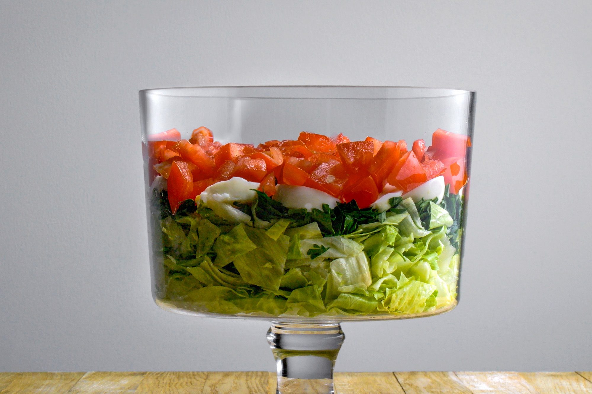 Table view shot of a clear glass bowl with layered salad, featuring a lettuce base topped with sliced eggs and diced tomatoes, on a wooden surface