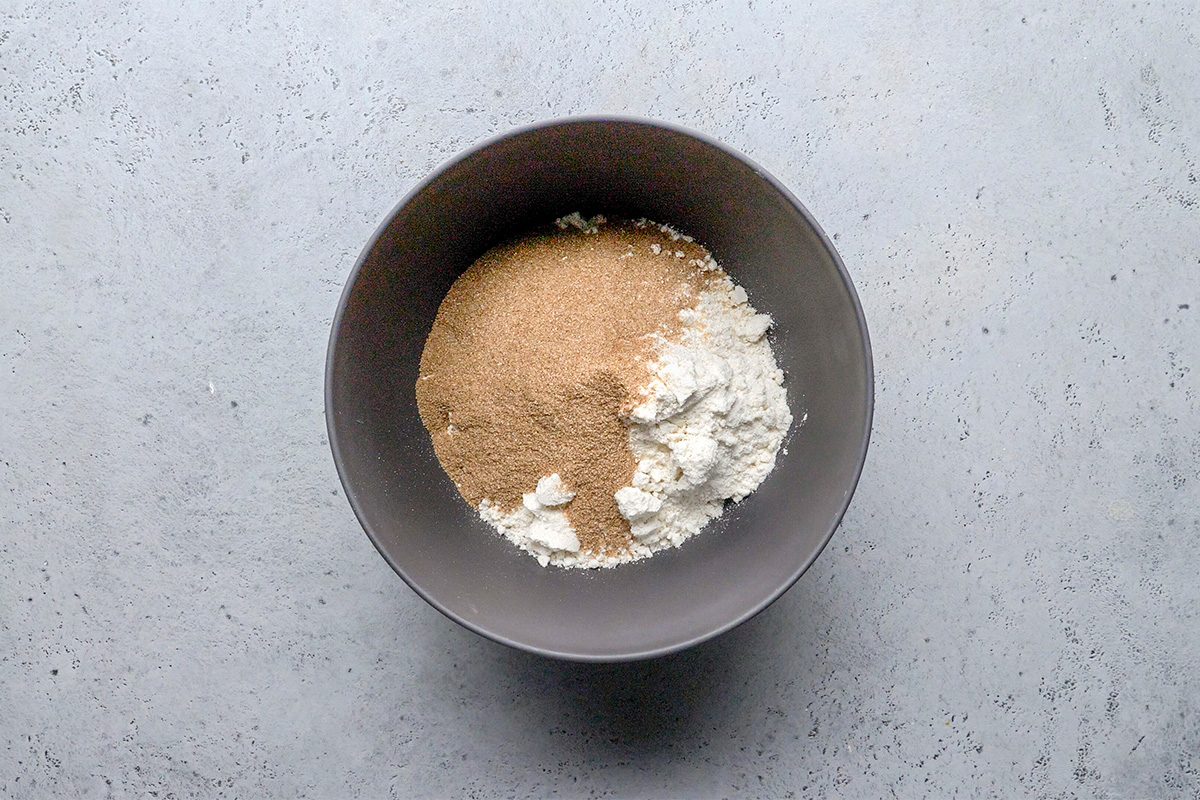 A gray bowl containing dry baking ingredients, including flour and brown sugar, sits on a light gray textured surface.