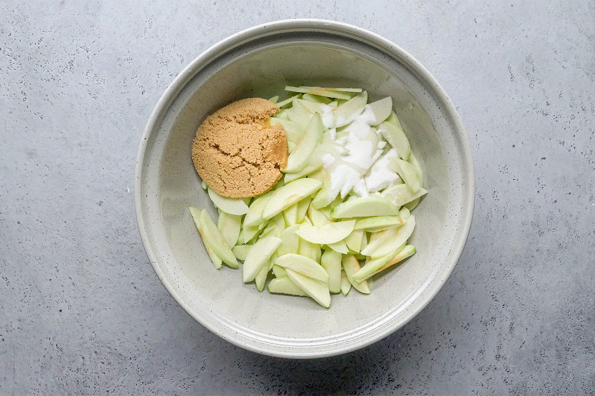 A bowl containing sliced green apples, a mound of brown sugar, and a pile of white granulated sugar, all placed on a light gray surface.