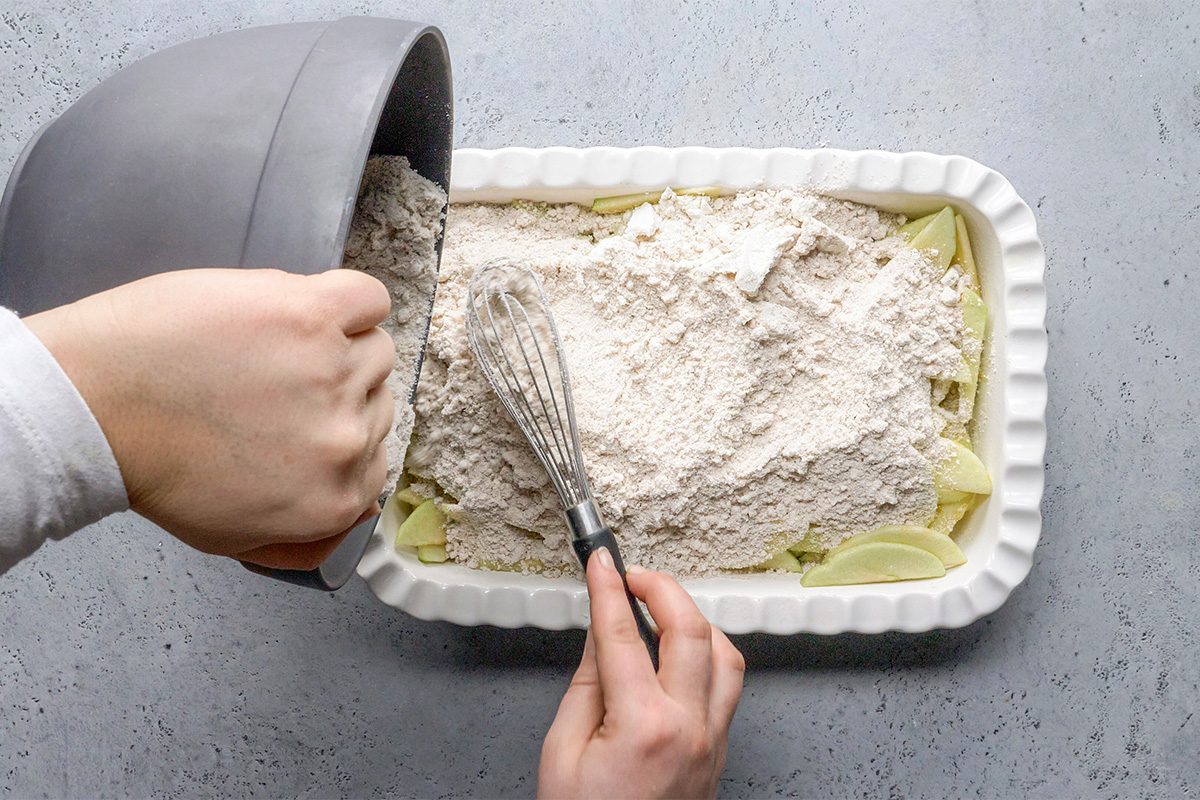 A person pours flour mixture from a gray bowl over sliced apples in a white baking dish while holding a whisk. The dish is on a light gray countertop.