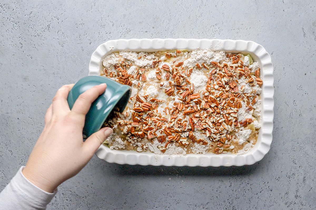 A hand pours a blue cup of liquid over an unbaked dessert in a rectangular white baking dish topped with chopped pecans and flour, set on a gray surface.