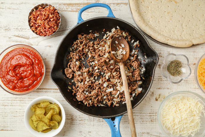 Top Shot of a skillet of cooked ground beef and onions is surrounded by pizza sauce; chopped bacon; shredded cheese; pickles; dried herbs; and an uncooked pizza crust on a wooden table background