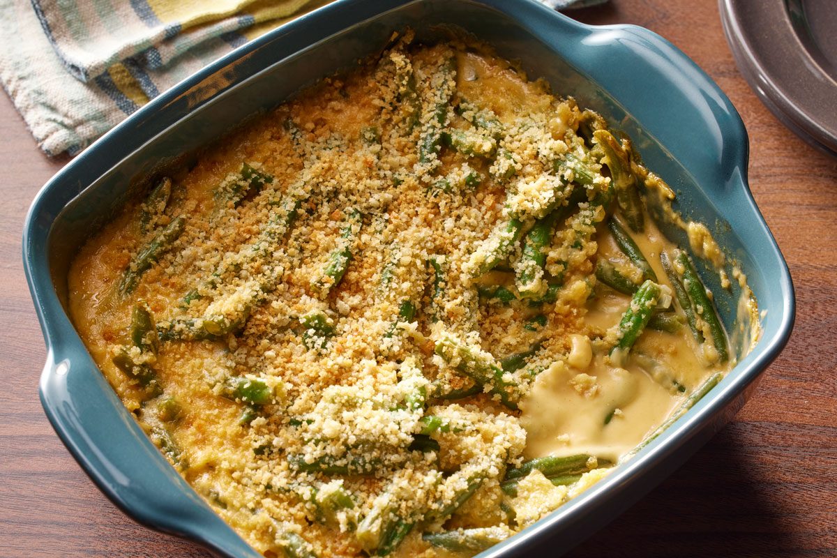 3/4th shot of A blue baking dish of Beer Cheese Green Bean Casserole topped with golden breadcrumbs, one corner scooped out, on a wooden table