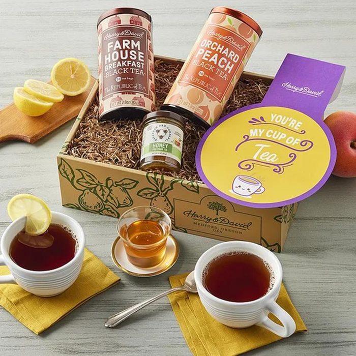 A gift box with two tins of tea, a jar of honey, a honey dipper, two cups of tea with lemon, and a paddle-shaped card that says 