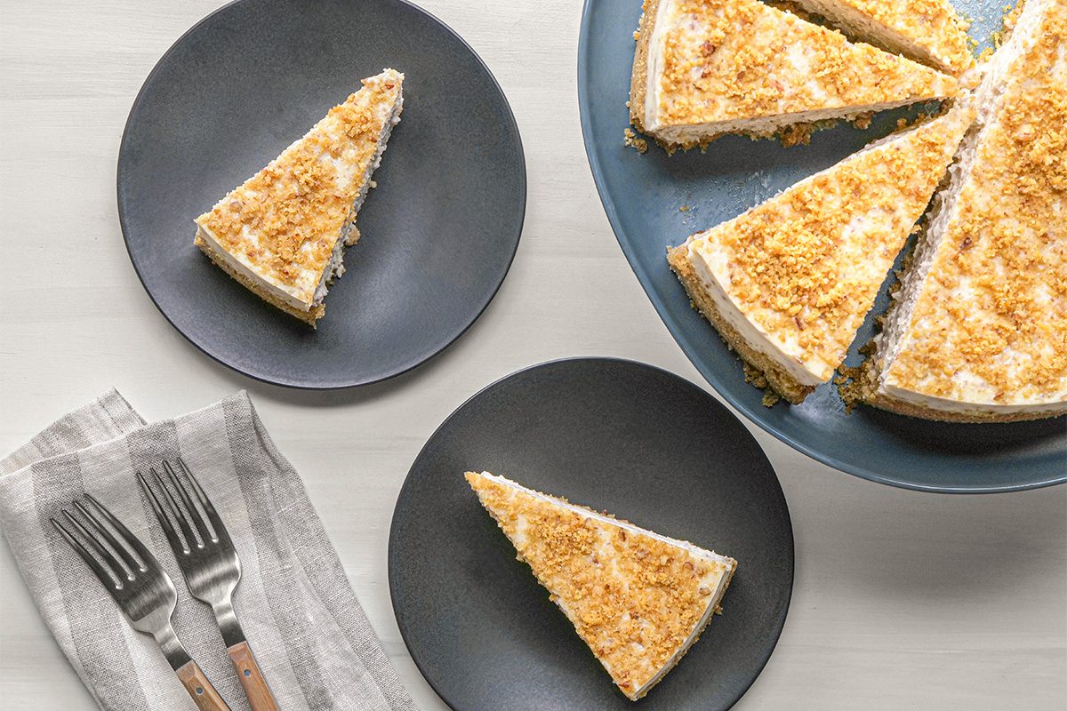 Two slices of crumb-topped cheesecake are served on black plates, with the rest of the cheesecake on a blue cake stand. Two forks rest on a folded napkin nearby, all set on a light-colored table.