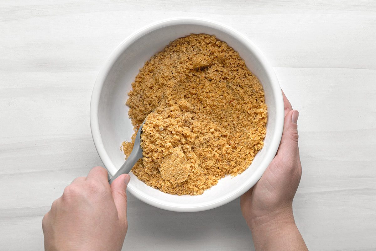 A person mixing crushed graham crackers in a white bowl with a spoon, viewed from above on a light-colored surface.