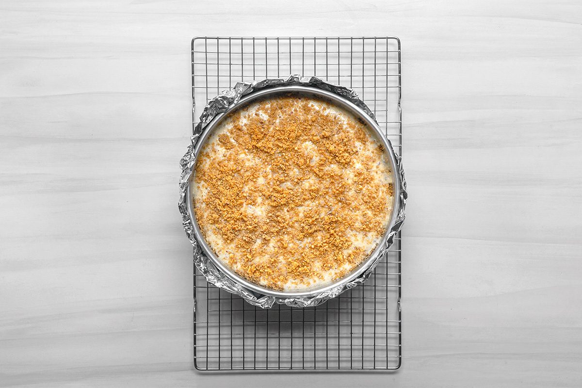 A round cheesecake in a foil-lined springform pan sits on a wire cooling rack. The cheesecake has a crumbly, golden topping and is viewed from above on a light-colored surface.
