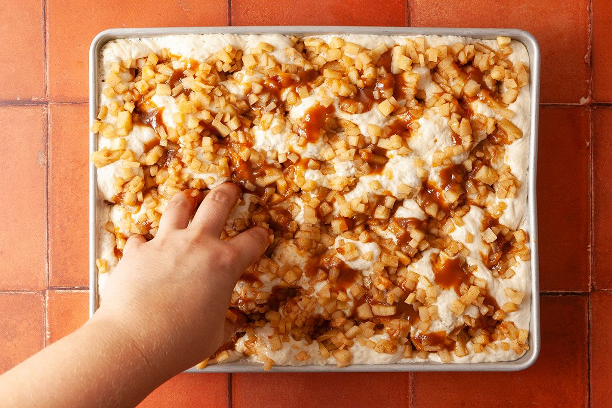 Overhead shot of a hand pressing into unbaked dough topped with diced apples and caramel sauce on a baking sheet, set on a terracotta tiled surface.