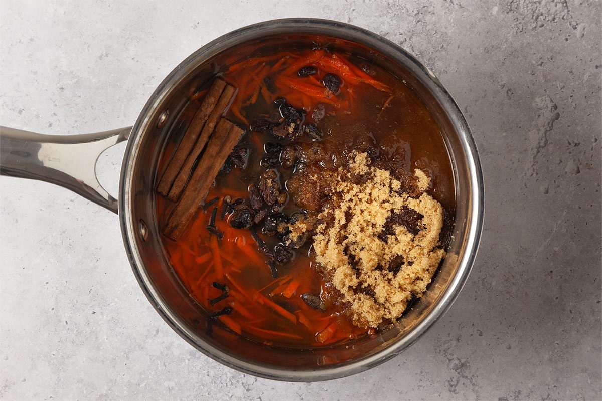 A saucepan filled with brown liquid, cinnamon sticks, star anise, orange peels, and a heap of brown sugar, sitting on a light gray surface.