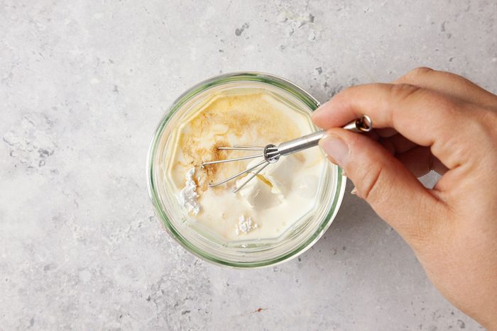 A hand holding a small whisk is mixing flour, yeast, and water in a glass bowl on a light-colored surface.